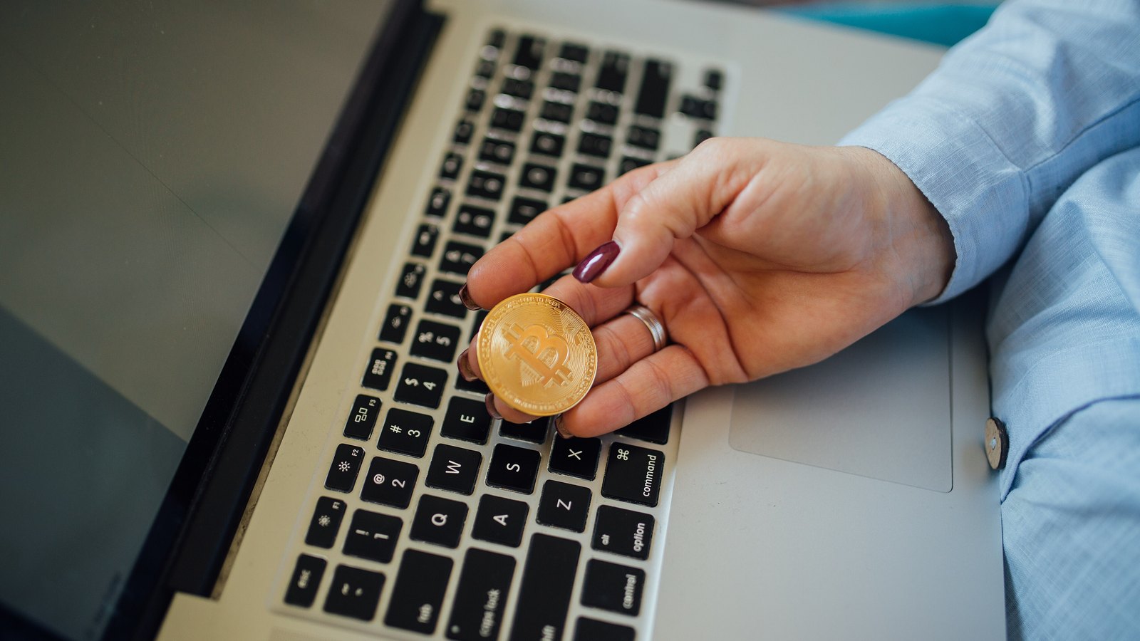 A physical bitcoin coin held in a hand in front of a laptop